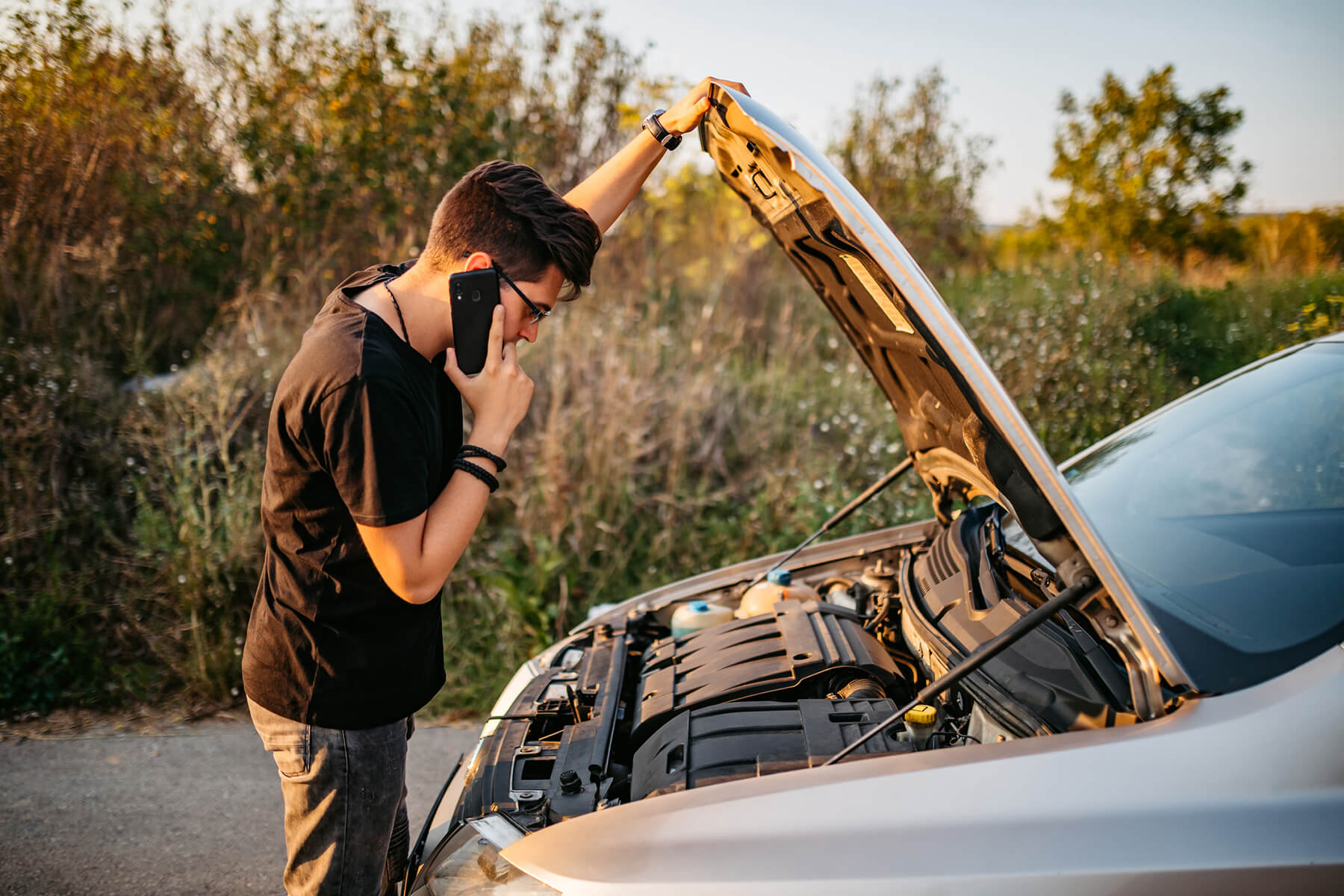 A young man looks under the hood of his car while making a call on his cellphone